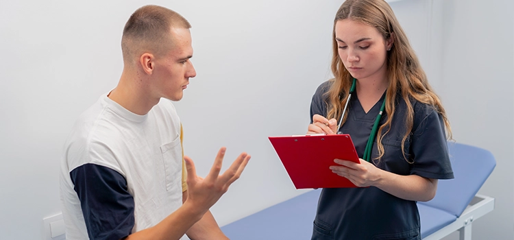 Healthcare assistant writing on a clipboard while listening to a male patient during a consultation in a medical room.