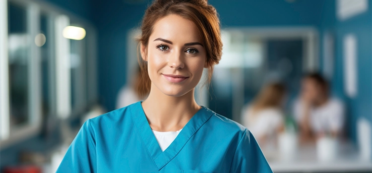 A smiling healthcare assistant in blue scrubs holding a clipboard in a medical facility.