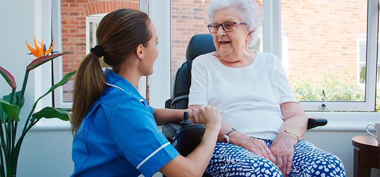 Healthcare assistant holding hands and chatting with a smiling elderly woman in a wheelchair indoors.